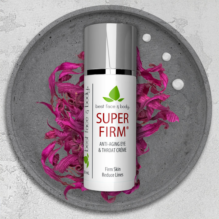 A jar of Super Firm Anti-Aging Eye & Throat Creme is displayed on a grey surface with decorative pink leaves around it.