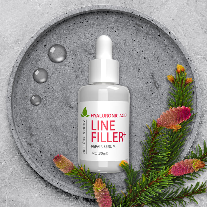 Hyaluronic Acid Line Filler Plus Repair Serum. White serum dropper bottle pictured overhead with stone background and greenery.