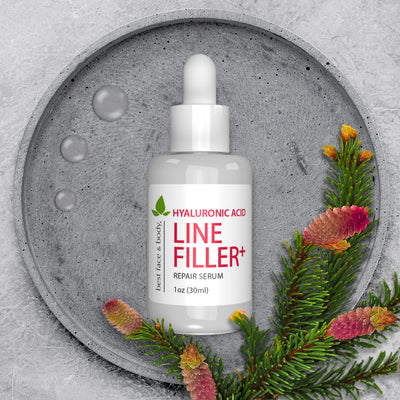 Hyaluronic Acid Line Filler Plus Repair Serum. White serum dropper bottle pictured overhead with stone background and greenery.