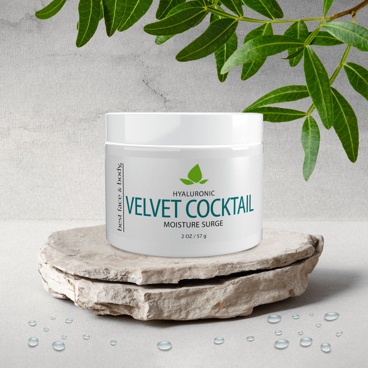 A white jar labeled 'Hyaluronic Velvet Cocktail Moisture Surge' is placed on a natural stone, with water droplets scattered around, and leaves in the background.