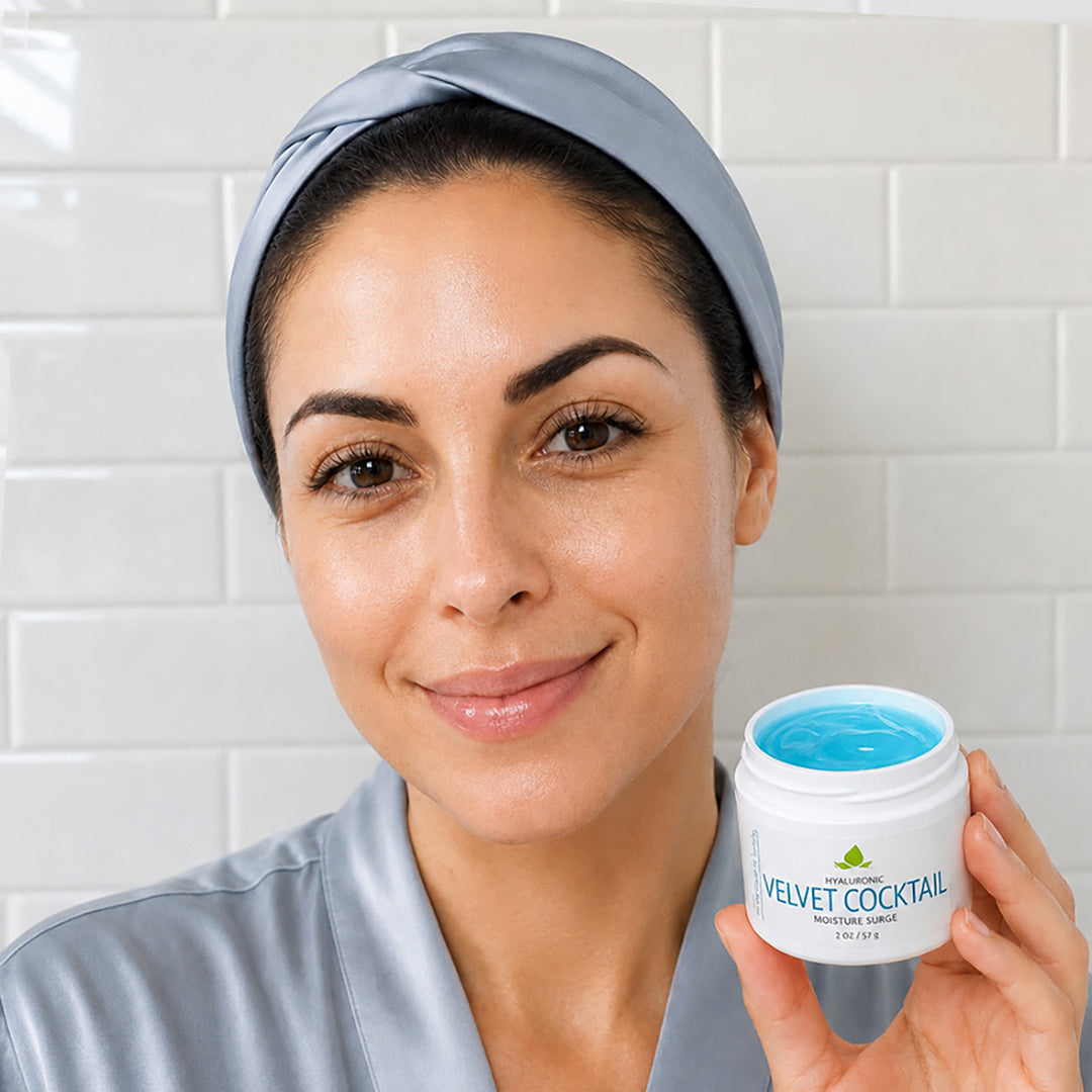 A woman wearing a light blue satin robe and matching headband holds up a white jar of Velvet Cocktail Hydrating Mask cream against a white subway tile wall background.