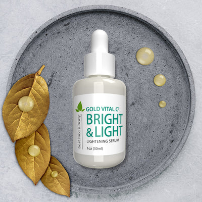 Gold Vital C3 Bright & Light Lightening Serum. 1 oz glass bottle with white dropper top, pictured on grey stone dish with decorative gold leaves and serum drops on the side.